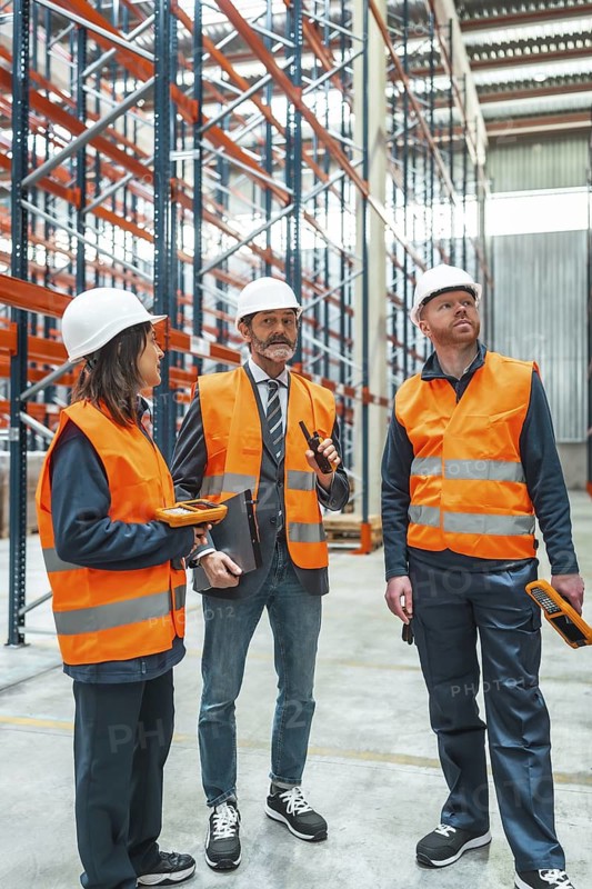 Warehouse workers and manager wearing safety vests and helmets, using walkie talkie and barcode scanner for inventory management and logistics operations