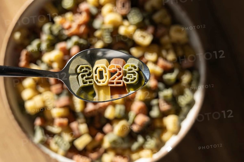 A spoonful of numerically shaped pasta displays the year 2026 above a bowl full of colorful number pasta, symbolizing new year celebrations