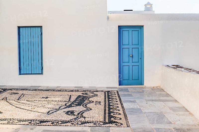 A serene Greek courtyard showcasing a blue door and window against a white wall in Plaka, Milos island. The mosaic patio adds texture to the minimalist aesthetic, capturing a calm Mediterranean vibe