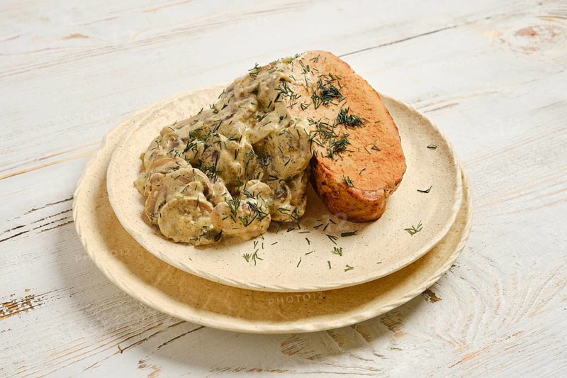 Creamy sauce with mushrooms covers slices of roasted pork meat. A sprinkle of herbs decorates the meal. Bright lighting shows details of the dish