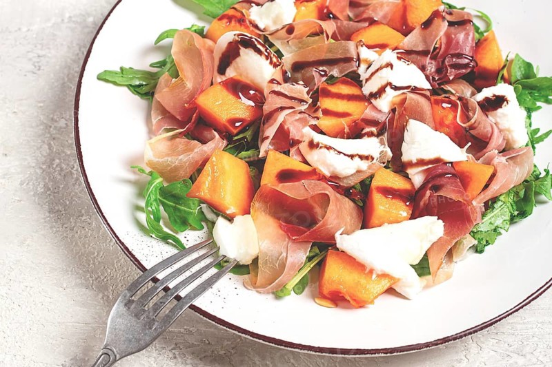 Salad with slices of ripe melon, prosciutto, mozzarella and fresh arugula, decorated with balsamic glaze