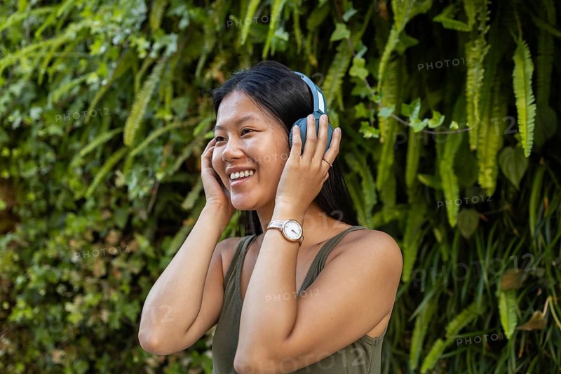 A cheerful Chinese woman wears headphones and enjoys music outdoors. Surrounded by lush greenery, she smiles brightly, embodying a relaxed and joyful summer city vibe