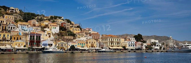 Colourful houses and boats line a tranquil coastline under a clear sky, Symi Town, Symi, Dodecanese, Greek Islands, Greece