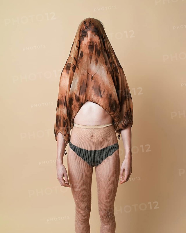 A young woman stands against a beige background in a studio, wearing a sheer top over her head while looking directly at the camera