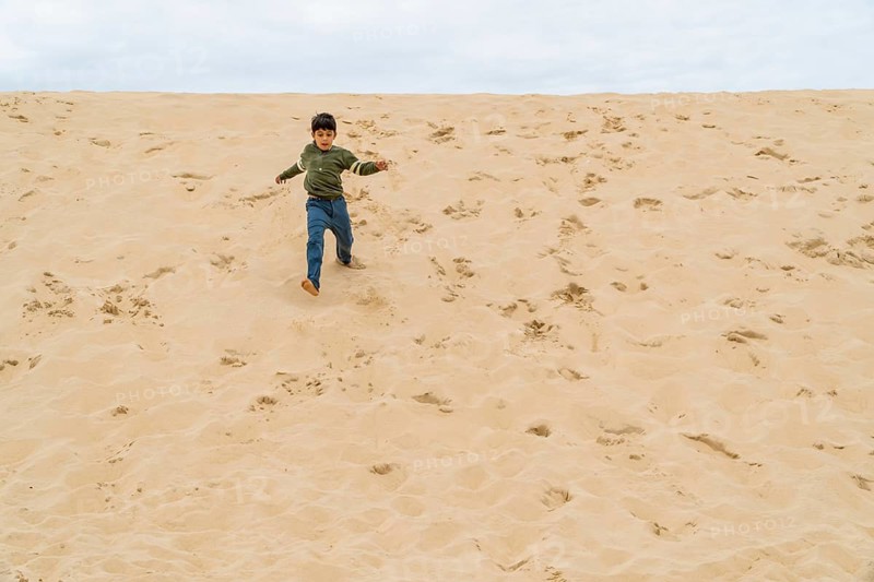 A young boy in casual clothing joyfully runs down the dune of Pilat under an overcast sky in France. The soft sand and wide open space convey freedom and adventure