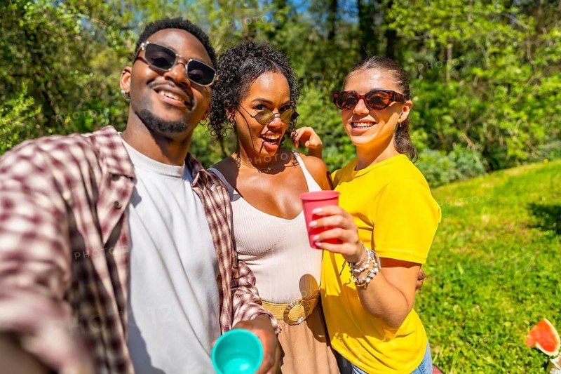 Three cheerful friends enjoying a picnic, taking a selfie and holding colorful cups on a sunny day in the park