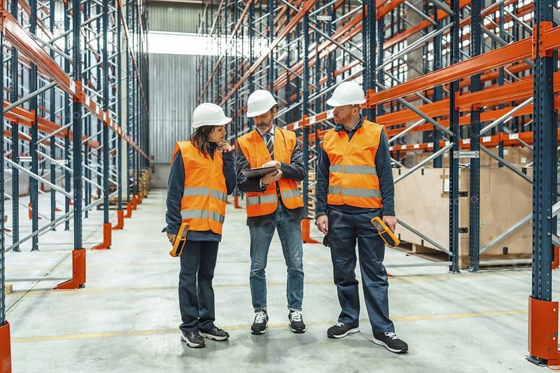 Warehouse workers and manager discussing logistics using a digital tablet, surrounded by shelves in a large distribution center