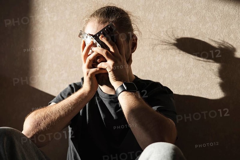 A man sits in a dimly lit room holding a blister pack of pills, deep in thought. The subtle shadow on the wall emphasizes his pensive mood and introspection