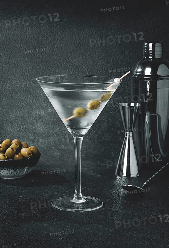 A drink is prepared in a bar setting. A martini glass is filled with clear liquid and has olives on a skewer. A cocktail shaker and a bowl of olives are nearby