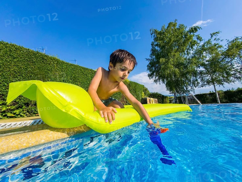 Young child enjoying a sunny summer day, dipping a water gun into the blue water from a brightly colored inflatable mattress at the side of a clean swimming pool