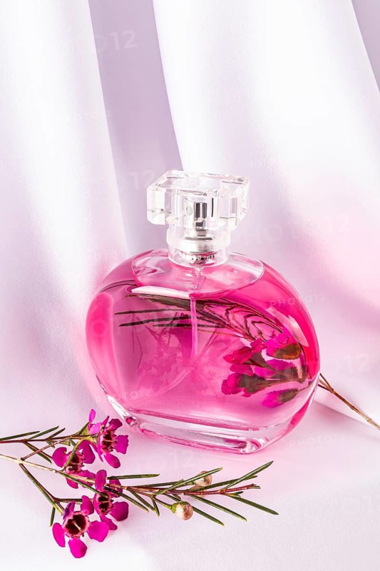 A sleek, pink-hued perfume bottle rests on elegant white fabric, surrounded by vibrant pink flowers. The image exudes luxury and a sense of floral elegance and freshness