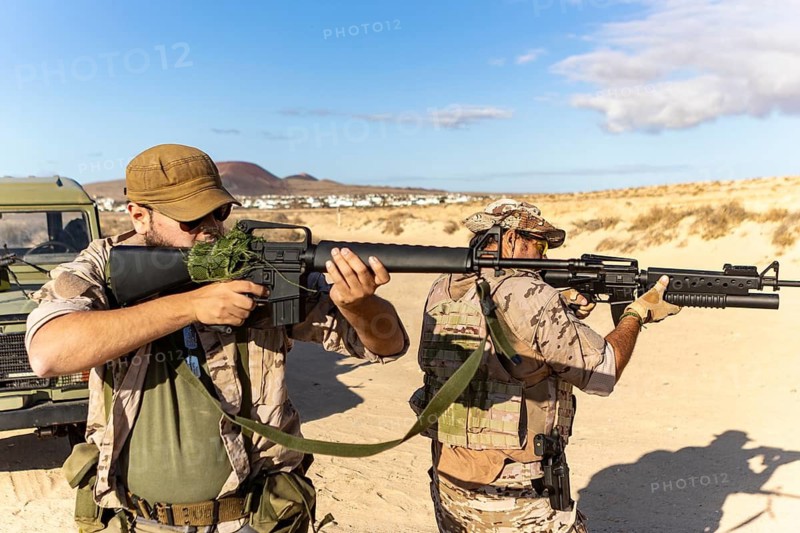 Two soldiers in camouflage gear participate in a military training exercise in a desert landscape. They hold rifles and stand next to a vehicle under a clear blue sky