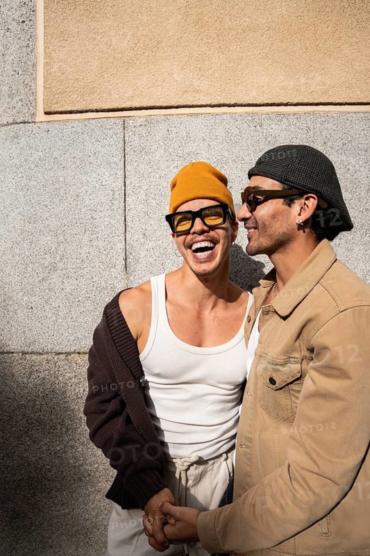 A gay couple joyfully embraces while holding hands outdoors. They wear stylish casual outfits and sunglasses, exuding happiness and love in the warm sunlight
