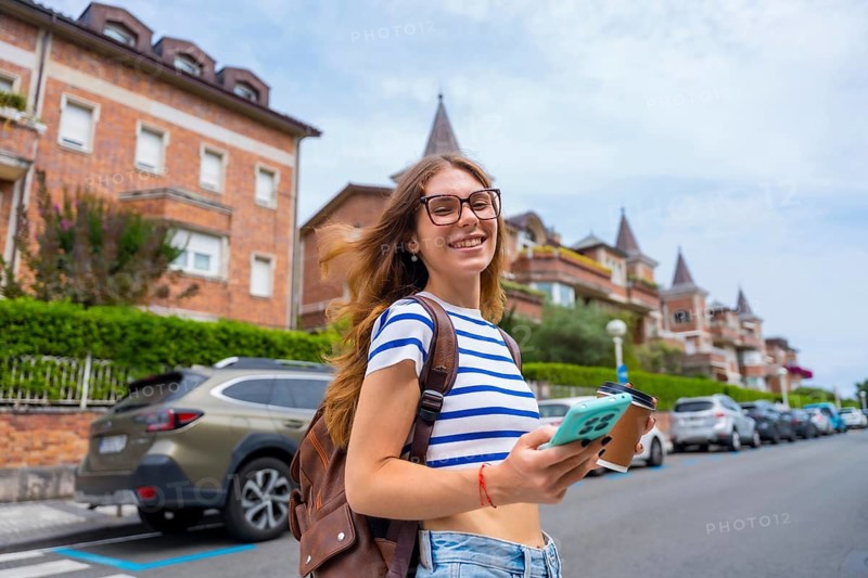 Young woman with glasses and backpack smiling while holding a smartphone and reusable coffee cup, walking along a sunny city street with buildings and parked cars