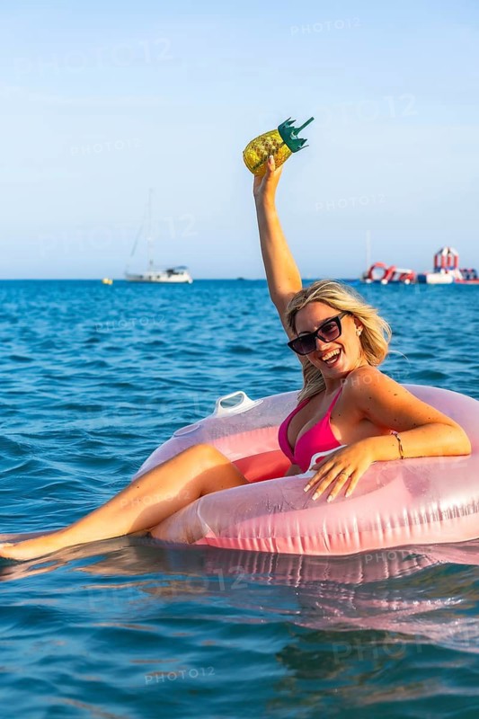 Young woman relaxing on an inflatable ring in the sea, holding a pineapple shaped drink while smiling and celebrating summer holidays on the beach