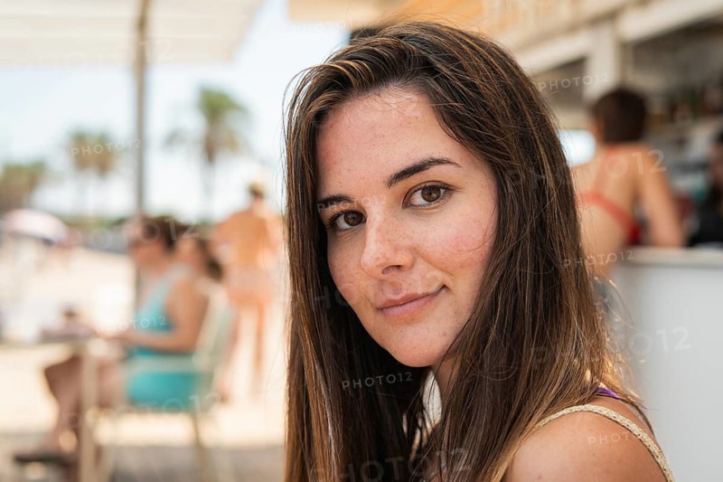 A woman with long hair and a relaxed expression enjoys her time at a beach side cafe. The blurred background shows people socializing under the sun, capturing a carefree summer vibe