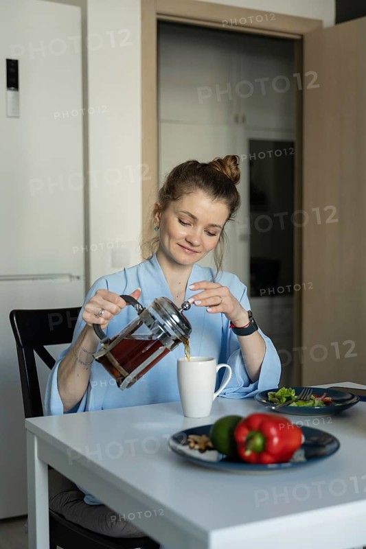 A woman in a casual setting pours tea into a mug at her kitchen table, creating a warm and cozy atmosphere. Fresh vegetables are seen on the table, enhancing the homely vibe