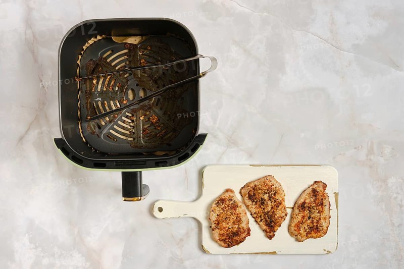Air fryer sits on counter with three cooked pork chops on white cutting board nearby. Kitchen setting shows clean surface and simple layout. Cooking equipment appears ready for use