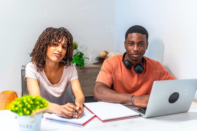 Two college students studying together at home, one using a laptop and the other taking notes