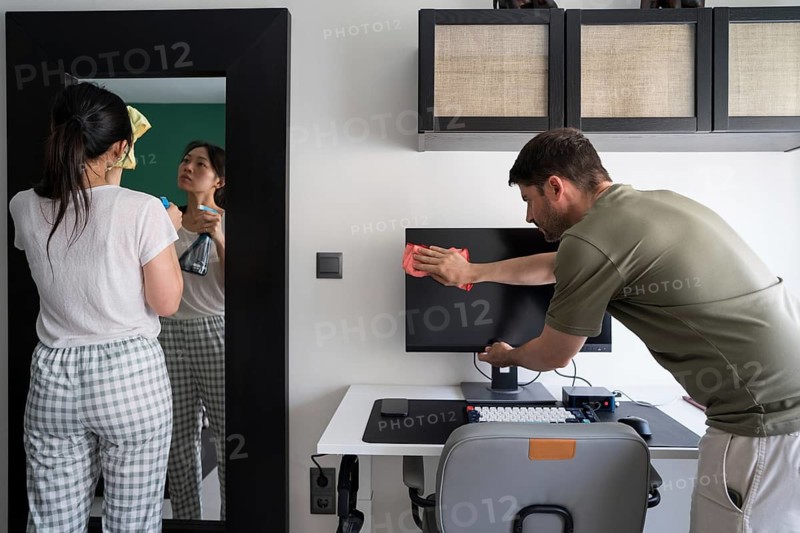 A contemporary couple engaged in household chores, exemplifying shared responsibilities. The woman cleans a mirror while the man wipes a computer screen, promoting cooperation and teamwork