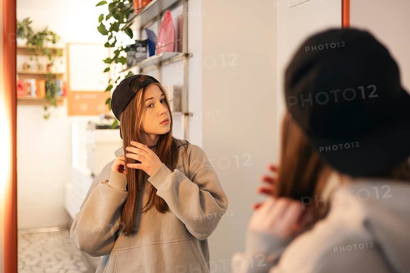 Teenage girl in a casual outfit adjusts her hair in front of a mirror. The room has modern decor and greenery, creating a cozy and inviting atmosphere