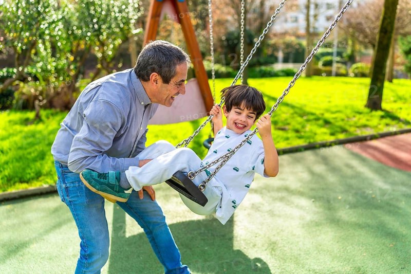 Grandfather and grandson enjoying a sunny day at the playground, pushing the swing and having fun