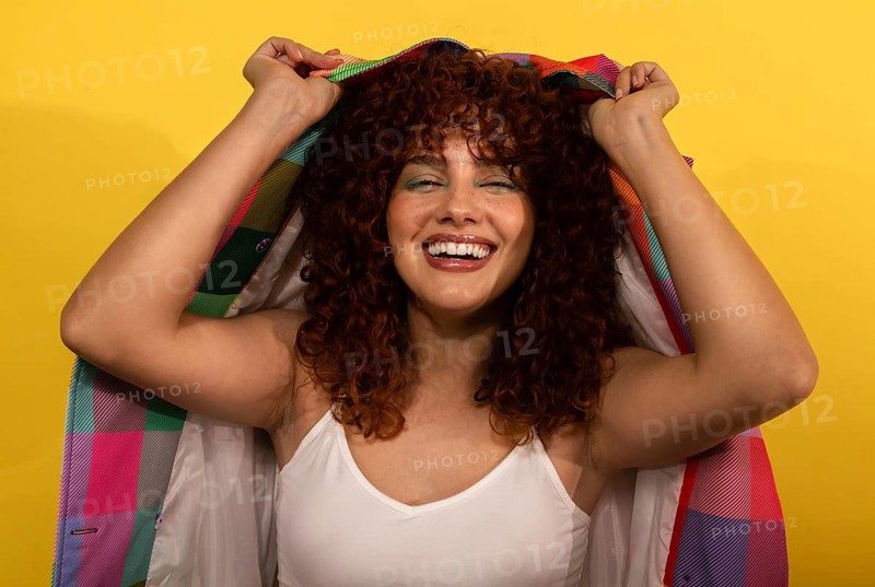 A woman joyfully smiles under a vibrantly colored fabric against a bright yellow background. She exudes happiness, showcasing her curly hair and wearing a white tank top