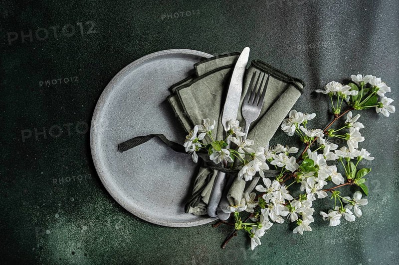 A minimalist clay plate with green napkin, silverware, and white blossoms on a dark textured surface. The arrangement evokes a fresh, springtime dining aesthetic