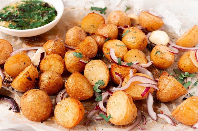 Roasted potatoes sit on a baking sheet next to chopped herbs and red onions. They are golden brown and ready to be served. Ingredients add color and flavor to the dish