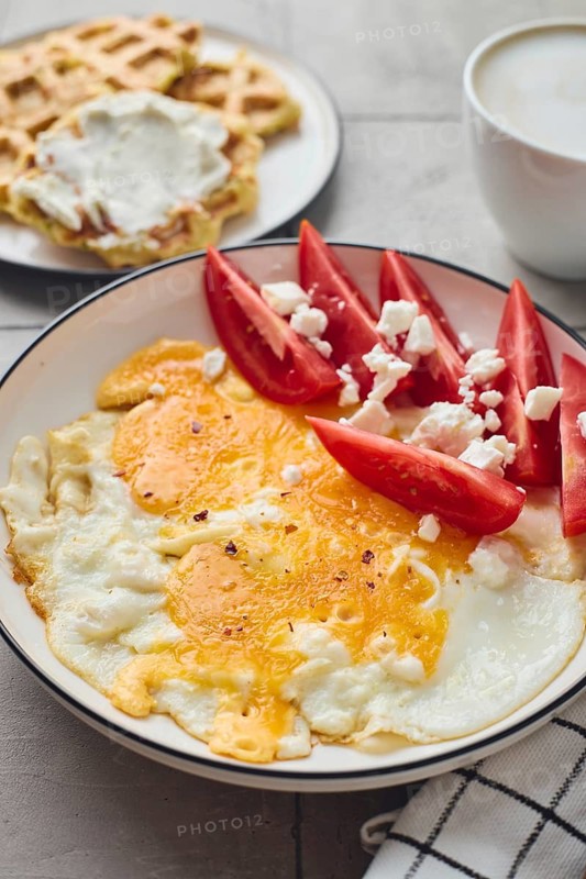 From above a mouth watering setup featuring a plate of fried egg topped with feta cheese, accompanied by fresh tomato slices and creamy Belgian waffles, served with a steaming cup of coffee