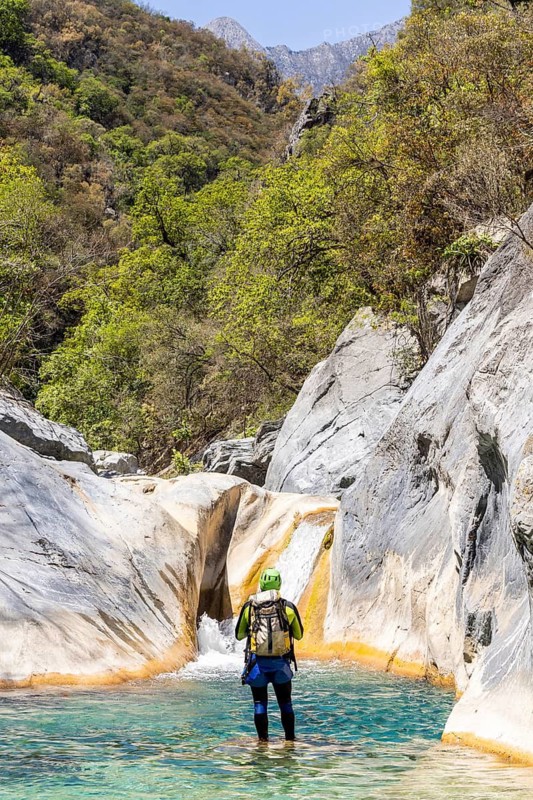 Back view of a canyoneer standing in a clear turquoise pool, preparing to climb a smooth natural slide among rocks in Matacanes, Nuevo Leon, Mexico. Adventure in nature