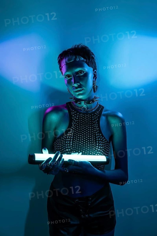 A futuristic cyberpunk woman stands confidently holding a glowing neon light against a vibrant blue backdrop. Her spiked accessories add an edgy, modern touch