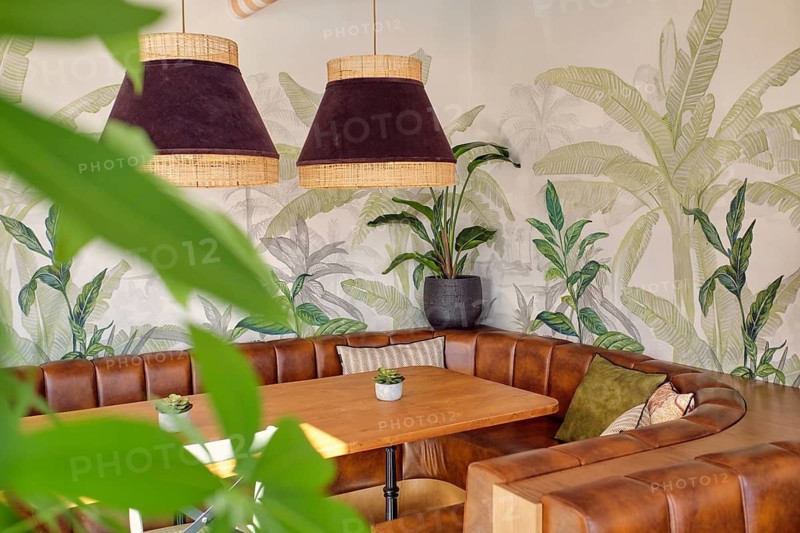A cozy corner of a restaurant featuring elegant leather booth seating, wooden tables, and tropical-themed wall decor accentuated by stylish pendant lights