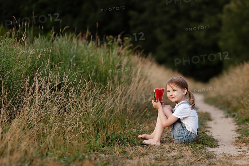 A child sits barefoot on a grassy path, enjoying a slice of juicy watermelon. The summer sun casts a warm glow, highlighting the lush greenery around them