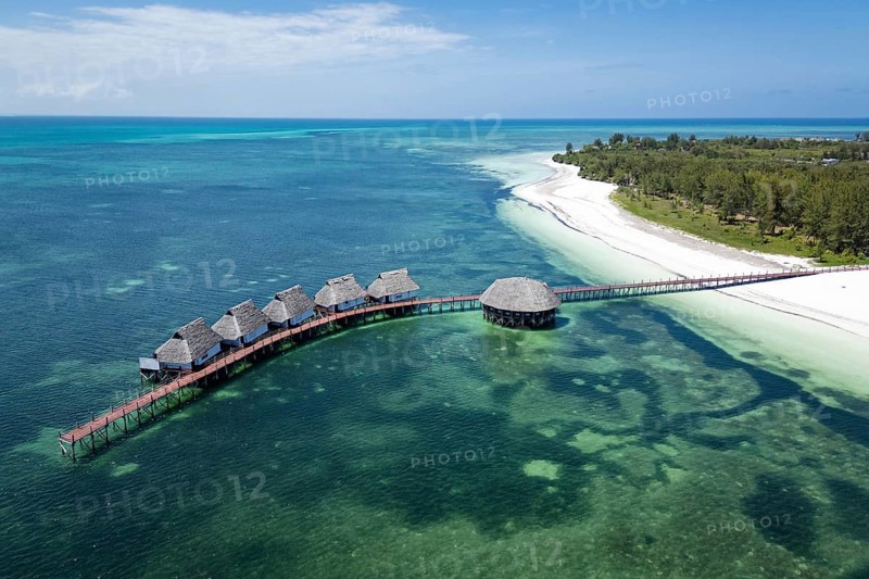 Aerial view of luxury overwater bungalows with thatched roofs and a wooden pier surrounded by emerald ocean and white sand beach in Zanzibar