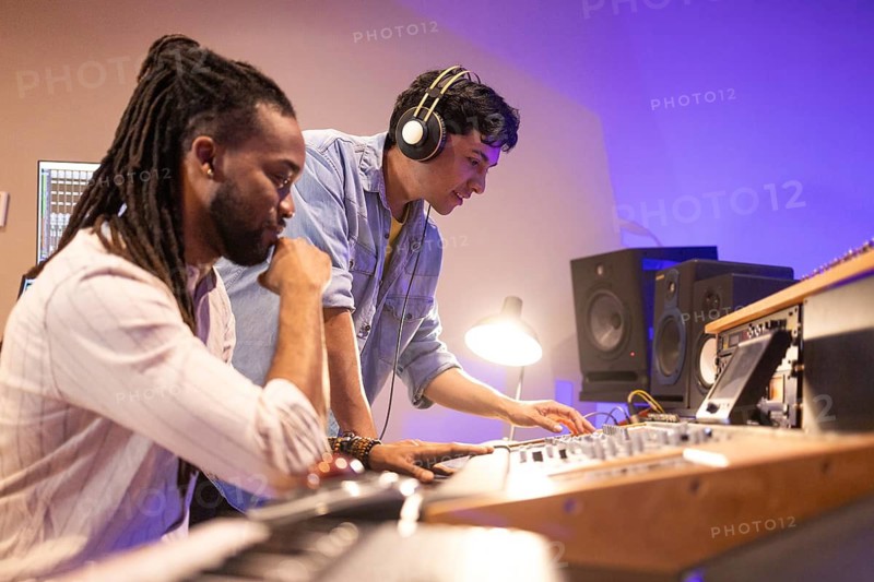 Two musicians collaborate in a state-of-the-art recording studio. One wears headphones, operating a soundboard, while the other observes, surrounded by audio equipment