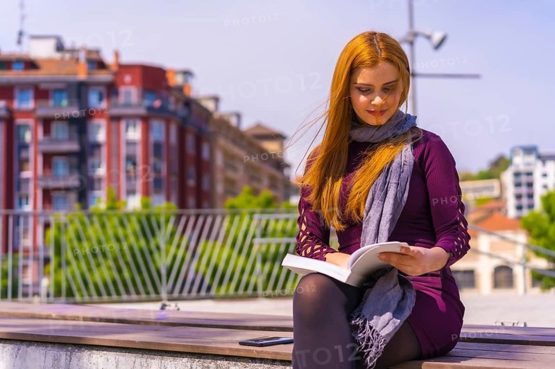 Beautiful woman in purple dress and scarf reading a book in a park in the city, imagining and enjoying reading