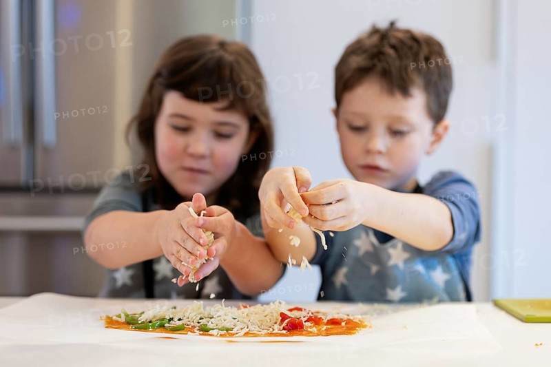 Two siblings are enthusiastically making a homemade pizza, sprinkling cheese over the base. They are focused and engaged, showcasing teamwork and creativity in the kitchen