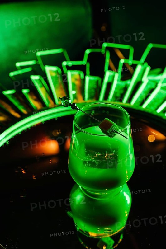A creamy vanilla gin cocktail with a citrus garnish, served over ice in a modern glass. The drink sits on a reflective tray, illuminated by vibrant green neon lighting for a futuristic feel