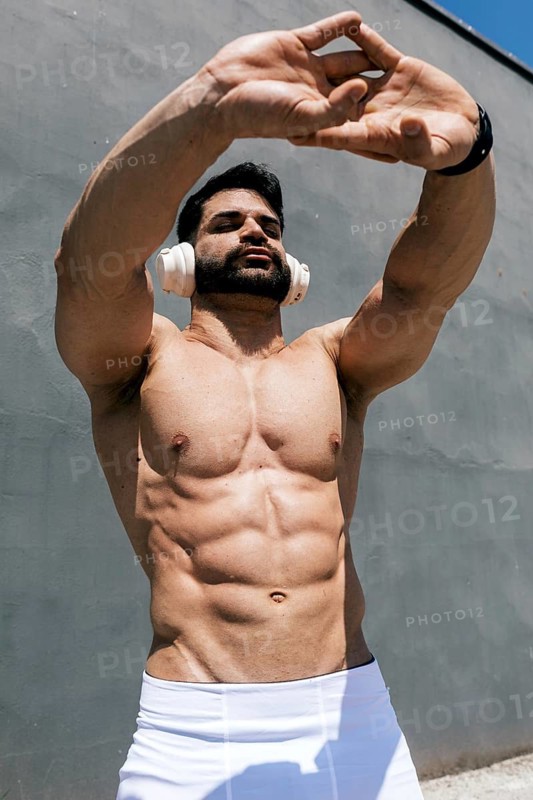 Muscular man exercising outdoors, wearing headphones, stretching under the sunlight. His toned physique highlights a dedication to fitness and a healthy lifestyle