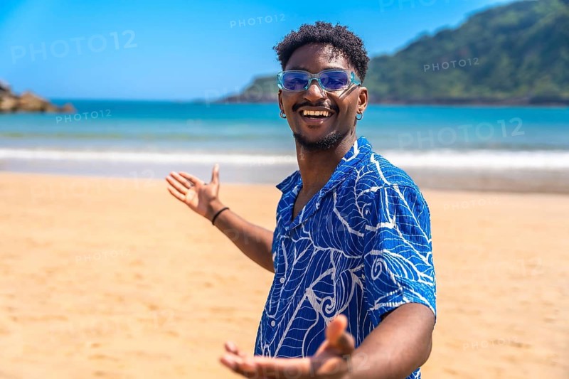 Young black man with sunglasses and hawaiian shirt enjoying summer holidays welcoming people to a tropical beach with open arms