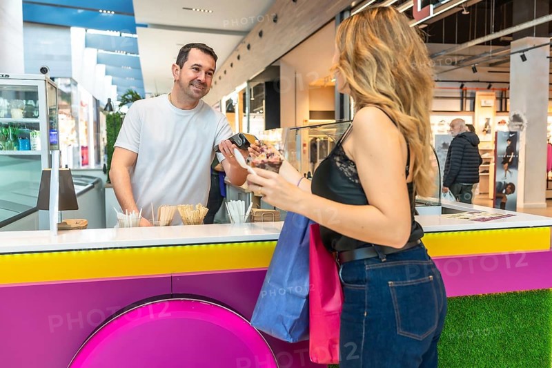 Smiling vendor processing a cashless payment for a customer purchasing ice cream, holding shopping bags, showcasing modern retail and consumer spending in a shopping center