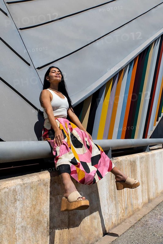 A woman in a vibrant, colorful outfit sits gracefully against a striking backdrop of modern architecture. The contrast highlights urban style and creative expression