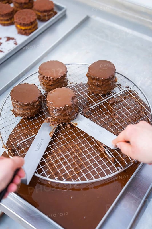 Two hands use knives to move several chocolate-glazed cakes on a drip net in the kitchen, vegan cake production, Haselstaller Hof, Gechingen, Germany