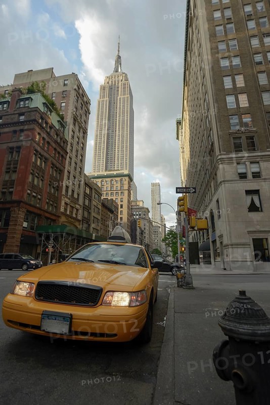 A classic yellow taxi is prominently featured in the foreground on a bustling Manhattan street, with the Empire State Building towering majestically in the background