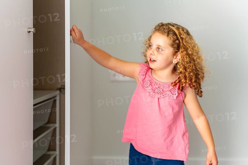 Young curly haired girl opens an empty wardrobe in her new room, exploring shelves and drawers with bright curiosity during a family move into their modern home