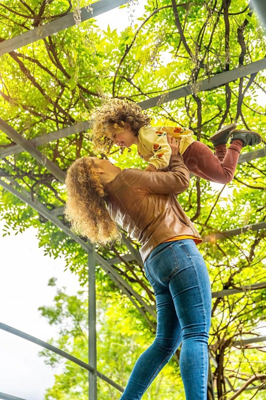 Vertical photo of a caucasuan blonde mother lifting playful girl standing on a park