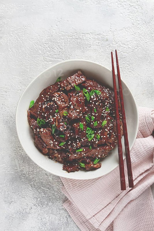 Crispy beef, with green onions and sesame seeds, in a bowl with chopsticks, on a textured surface, on a light background, no people