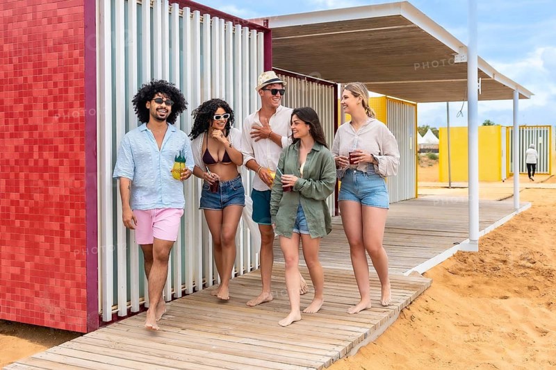 Group of diverse friends walking barefoot on a wooden boardwalk, smiling and holding refreshing drinks on a sunny day at a beach resort with colorful cabins