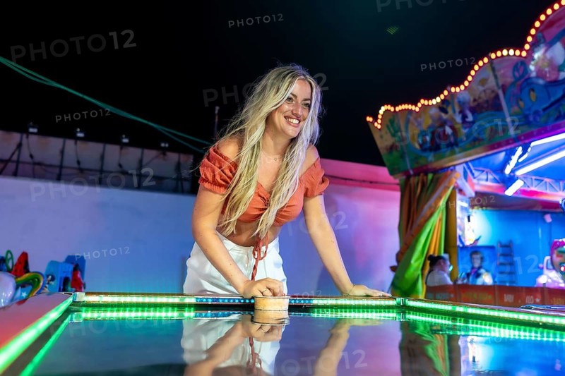 Young blonde woman smiling, playing air hockey at a vibrant night carnival, with colorful neon lights reflecting off the game table, creating a joyful, dynamic atmosphere of summer fun